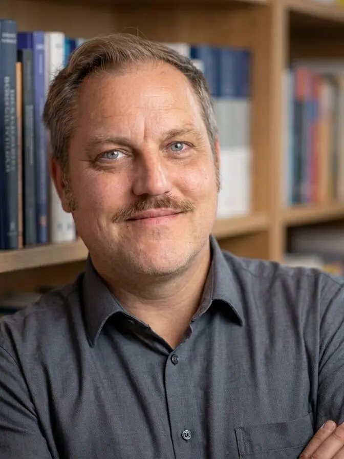 Heiner Bäumer, an ein Bücherregal gelehnt, Arme verschränkt, dezentes Lächeln. Szene KI-inszeniert auf Basis einer echten Aufnahme.